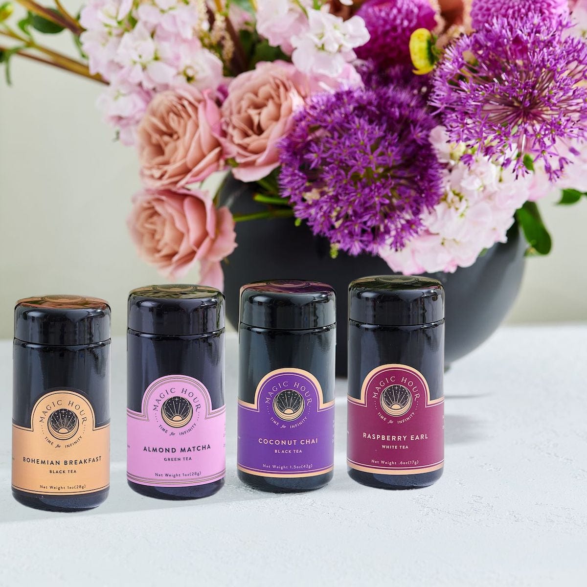 Four **Luxury Traveler Gift Box** sets from **Magic Hour** are arranged in front of a large black vase filled with pink, purple, and white flowers. The scene is set on a light-colored surface.