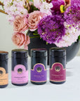 Four **Luxury Traveler Gift Box** sets from **Magic Hour** are arranged in front of a large black vase filled with pink, purple, and white flowers. The scene is set on a light-colored surface.