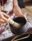 A person is using a wooden whisk to froth Magic Hour Tea in an Alibaba Ceremonial Tea Bowl. The individual is wearing a patterned garment and a gold ring, with part of a gold necklace visible. A tray with other utensils is in the background.