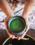 A person holding a black ceramic Ceremonial Tea Bowl filled with vibrant green Magic Hour Tea matcha. The person is seated and wearing a patterned garment with hues of red and beige. The background features a wooden surface and partial view of another black bowl from Alibaba.