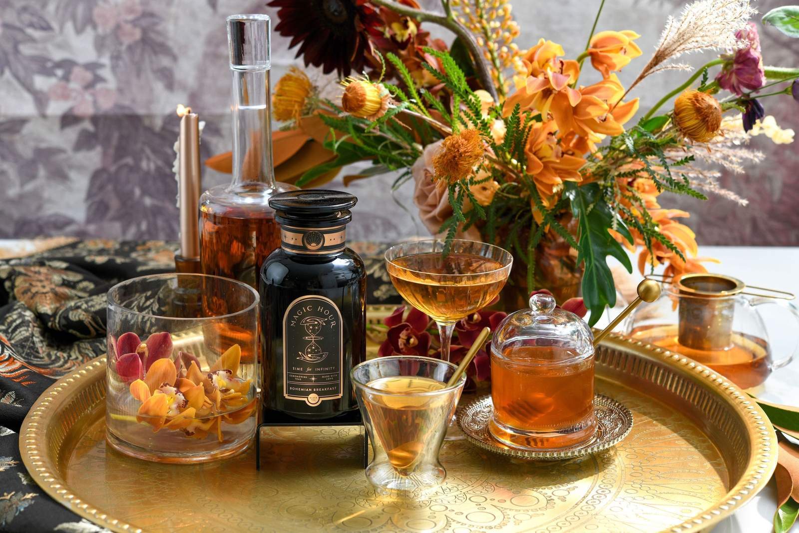A golden tray holds an assortment of drinks in fancy glassware, a black bottle, and a honey jar. Vibrant orange and yellow flowers in a bouquet add a touch of elegance, with a fabric featuring intricate patterns underneath. Candles are also present in the background, alongside Magic Hour's Tea Connoisseur Collection.