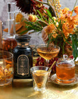 A golden tray holds an assortment of drinks in fancy glassware, a black bottle, and a honey jar. Vibrant orange and yellow flowers in a bouquet add a touch of elegance, with a fabric featuring intricate patterns underneath. Candles are also present in the background, alongside Magic Hour's Tea Connoisseur Collection.