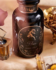 A glass bottle labeled "Magic Hour Astrology Bundle: Aries, Taurus, Gemini" is centered on a table adorned with various items. The label features an illustration of an archer with celestial designs. Surrounding the bottle are a purple flower, a golden dish with dried herbs, and a glass perfume bottle of Magic Hour Tea.