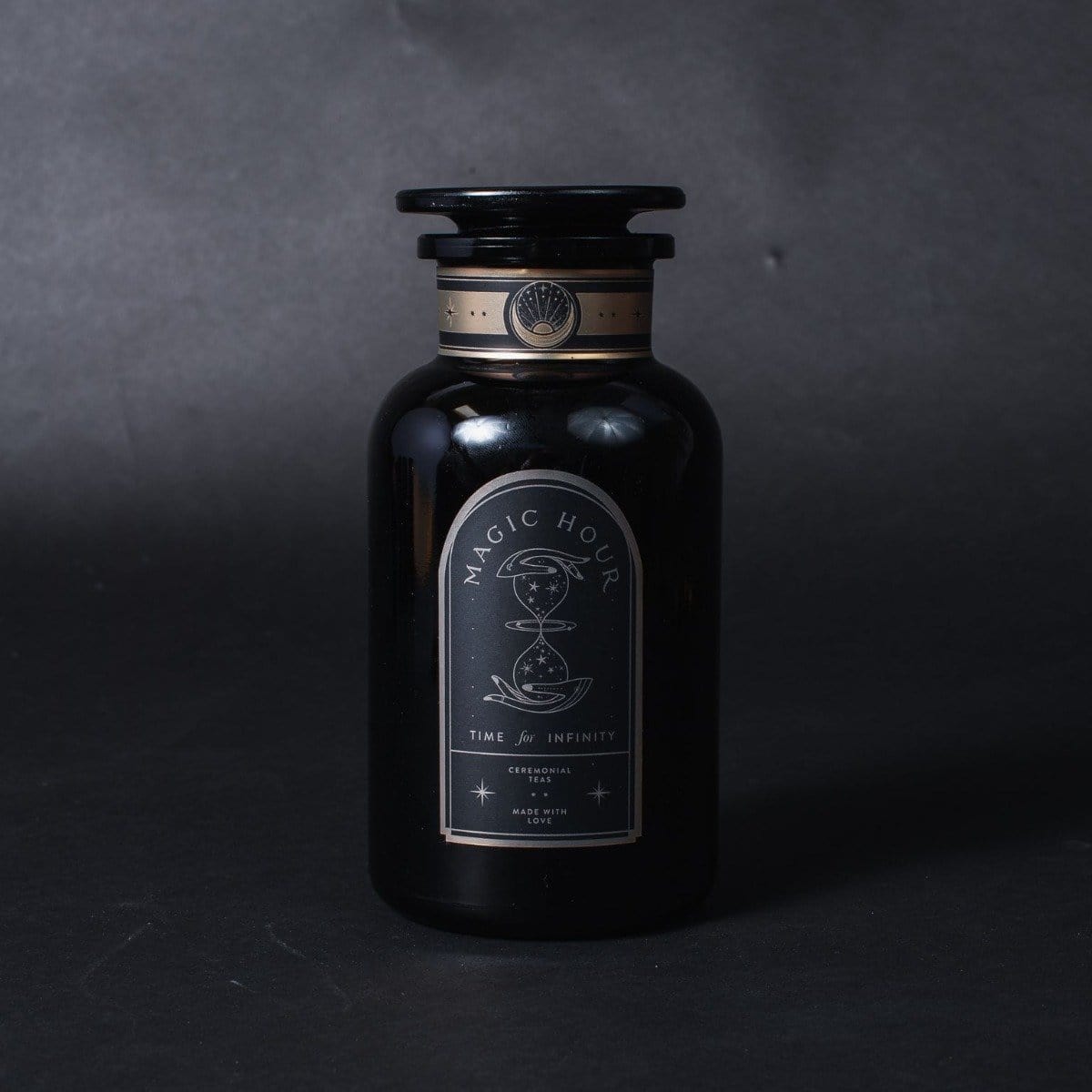 A dark glass bottle with a black label featuring the words "Bliss: Sacred Tulsi Adaptogen Tea" and an illustration of an hourglass. The label also reads "Time & Infinity," hinting at the exceptional quality of Magic Hour, and has additional decorative elements. The bottle is set against a dark background.