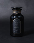 A dark glass bottle with a black label featuring the words "Bliss: Sacred Tulsi Adaptogen Tea" and an illustration of an hourglass. The label also reads "Time & Infinity," hinting at the exceptional quality of Magic Hour, and has additional decorative elements. The bottle is set against a dark background.