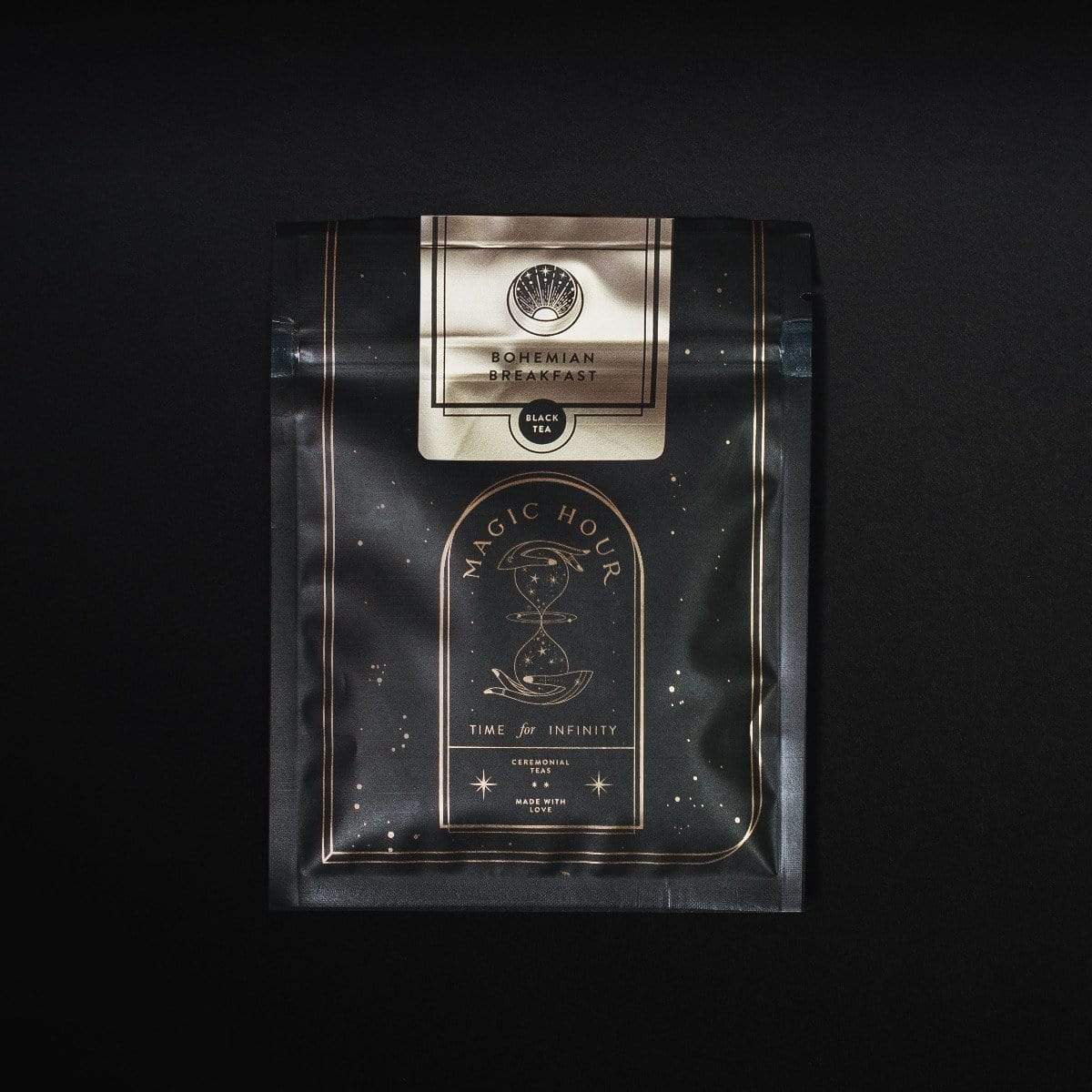 A sleek black package of "Sip Magic: Signature Blends of Magic Hour Ritual Gift" featuring gold accents and a central hourglass emblem, with the label "Time for Infinity" below it. This Organic Loose Leaf Tea by Magic Hour boasts a sophisticated and mystical design set against a dark background.