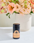 A small dark-colored jar with a beige label stands on a white surface in front of a vase filled with pink and peach flowers. The product label reads "Bohemian Breakfast™ Violet Glass Traveler." This organic tea by Magic Hour exudes freshness, enhanced by the brightly lit, airy atmosphere.