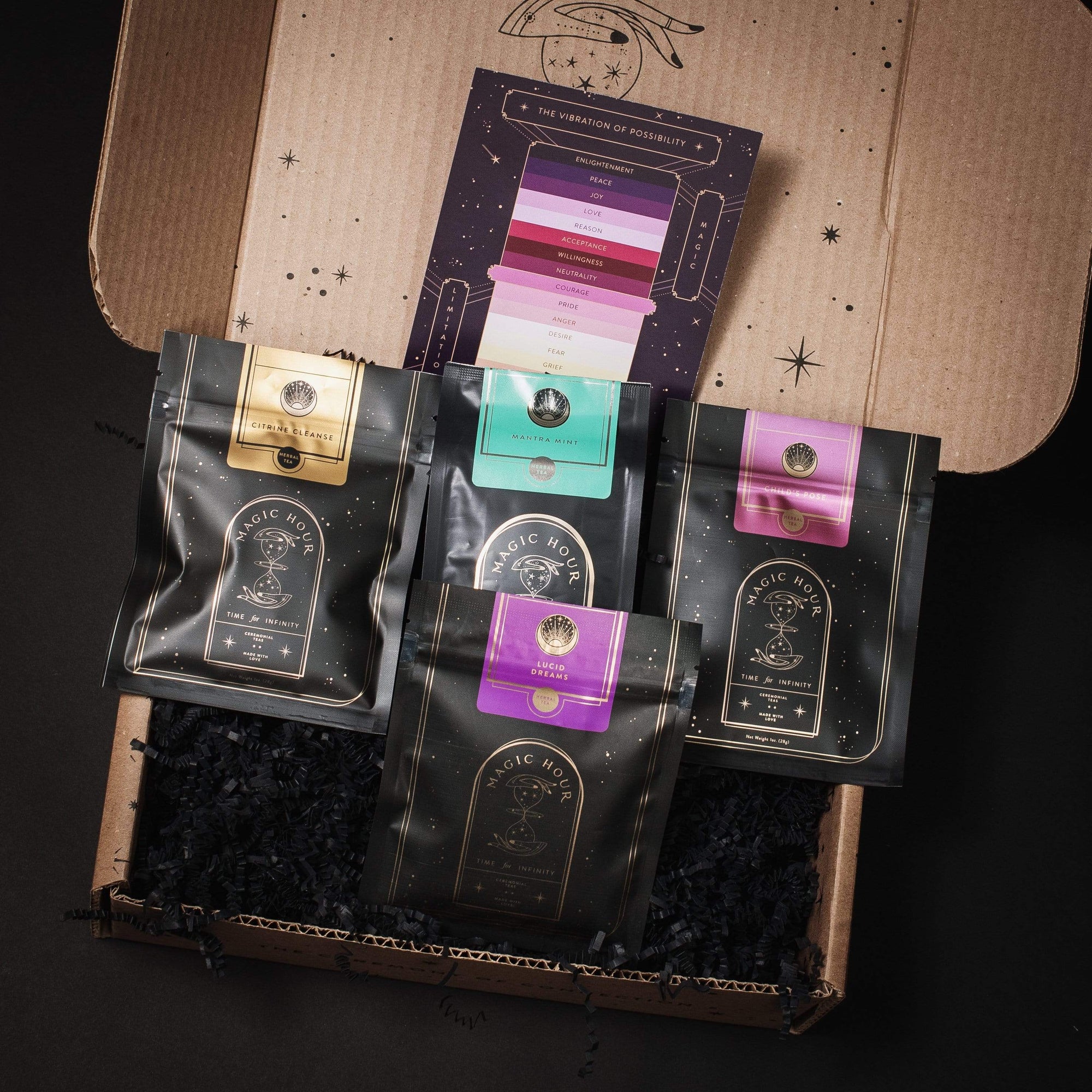 A gift box contains four black packages with colored labels arranged neatly inside. Each package features a design including the words "Club Magic Hour." Behind the packages, there is a colorful card titled "The Vibration of Possibility." The box and card have a starry design, perfect for an organic tea lover.