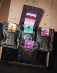 A gift box contains four black packages with colored labels arranged neatly inside. Each package features a design including the words "Club Magic Hour." Behind the packages, there is a colorful card titled "The Vibration of Possibility." The box and card have a starry design, perfect for an organic tea lover.