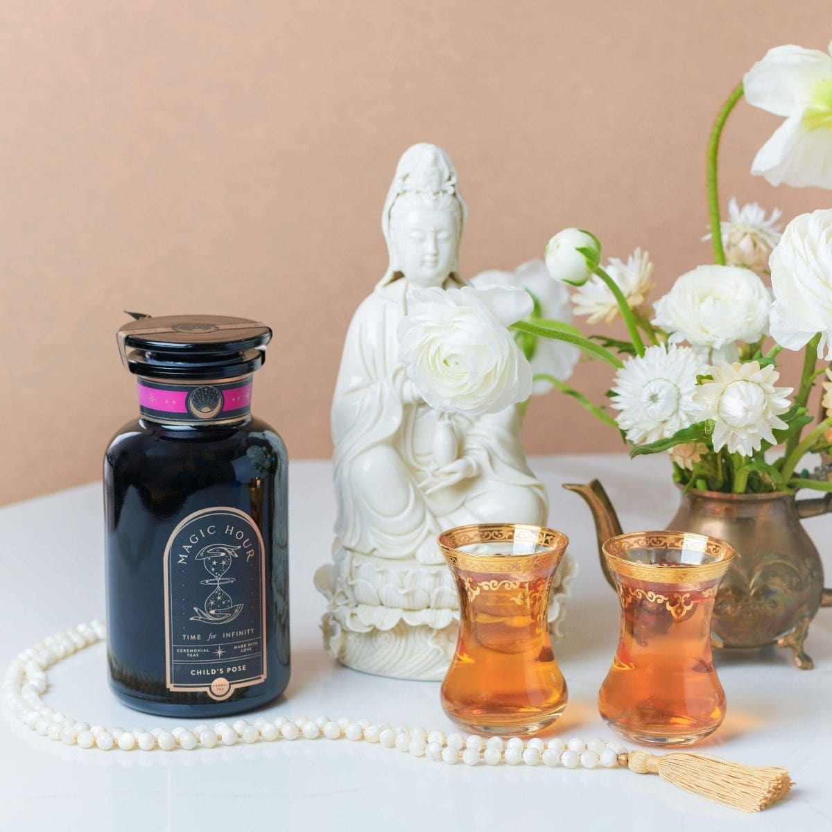 A still life image featuring a jar of tea from the Self-Love Box With Child's Pose, two golden-rimmed glasses filled with Child's Pose Herbal Tea, a white statue of a serene figure among white flowers, a string of white beads, and a small ornate brass teapot arranged on a white table with a neutral background.