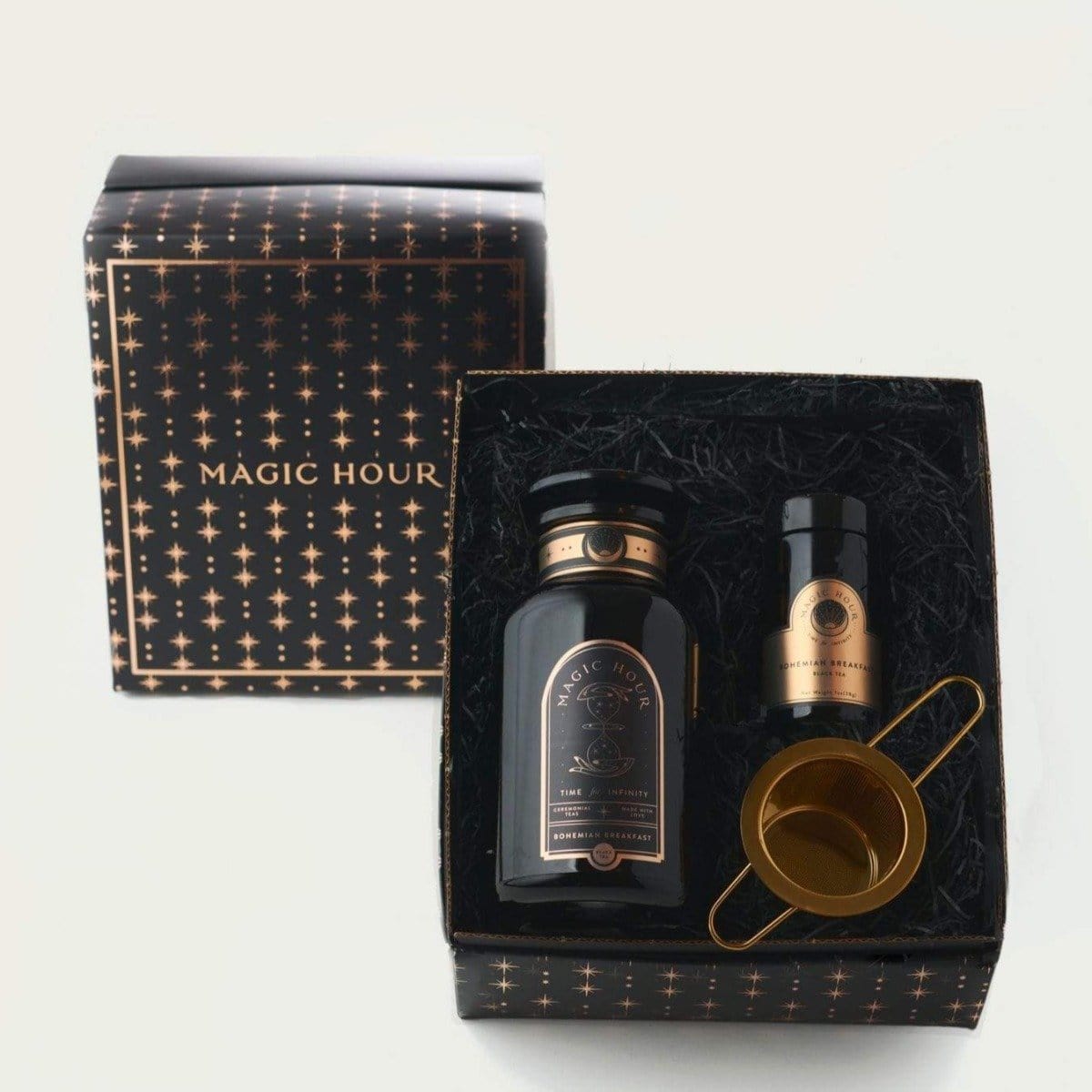 A gift set in a black and gold star-patterned box labeled "Magic Hour Bohemian Breakfast Apothecary & Traveler Set with Midas Touch," containing a black bottle, a small gold-capped vial, and a gold strainer. The box is open, showcasing the items carefully arranged on a bed of black crinkle paper.