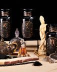An arrangement of decorative items including three black glass jars labeled "Aries," "Taurus," and "Gemini." The scene features a brown teapot filled with Astrology Bundle: Aries, Taurus, Gemini - Magic Hour by Magic Hour, two glass perfume bottles, a chess piece-like statue, and various trinkets on a beige surface with a black background.
