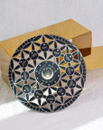A round black and silver decorative plate with intricate geometric patterns and text "Crystal Grid: Flower of Life Tea Ceremony Altar" at the center, surrounded by smaller circles with text like "gratitude," "immunity," and "healing." The plate is set against a white cloth backdrop, next to a golden rectangular object.