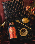 A luxurious tea gift set titled "Season of Abundance Gift Set" by Magic Hour lies open on a dark fabric. The set includes a tin of loose leaf tea with a red and gold label, a gold tea strainer, and a gold stirring spoon. Nearby is a golden teapot and a bouquet of flowers.