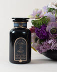 A black bottle labeled "Magic Hour - Earl Greater Grey: Tea for the Bright & Bold" sits beside a vibrant bouquet of purple and white flowers. Featuring an apothecary design with a cosmic hourglass label, this mysterious vessel contains Magic Hour's finest loose leaf tea, and the flowers are arranged in a black vase.