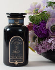 A black bottle labeled "Magic Hour - Earl Greater Grey: Tea for the Bright & Bold" sits beside a vibrant bouquet of purple and white flowers. Featuring an apothecary design with a cosmic hourglass label, this mysterious vessel contains Magic Hour's finest loose leaf tea, and the flowers are arranged in a black vase.