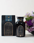 A black bag and a black jar of Magic Hour's "Earl Greater Grey: Tea for the Bright & Bold" loose leaf tea are placed next to a vase containing a bouquet of flowers with purple, white, and pink blooms. The packaging, which highlights the organic tea blend, features gold and white text with a mystical design.