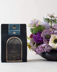 A black package of Magic Hour organic tea sits on a white surface next to a bouquet of purple and white flowers in a black vase. The tea package has a label that reads "Earl Greater Grey: Tea for the Bright & Bold" and "Time & Infinity" with an hourglass graphic, making this loose leaf tea experience timeless.