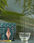 A geometric-patterned teal and pink box of Matcha 1111: Highest Grade of Ceremonial Matcha by Magic Hour sits next to a transparent glass bowl on a table. A bamboo scoop with green powder lies in the foreground, hinting at the organic loose leaf tea inside. The background has glass panels with vertical ridges and hanging green ferns.