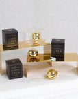 A display featuring two black boxes labeled "Sip Royaltea Gift Set," a glass cup, and three golden tea infusers with perforated spherical shapes and attached chains. Two infusers are opened showing the inside. The setup elegantly showcases the Magic Hour experience, perfect for steeping organic loose leaf tea on golden stands against a white backdrop.