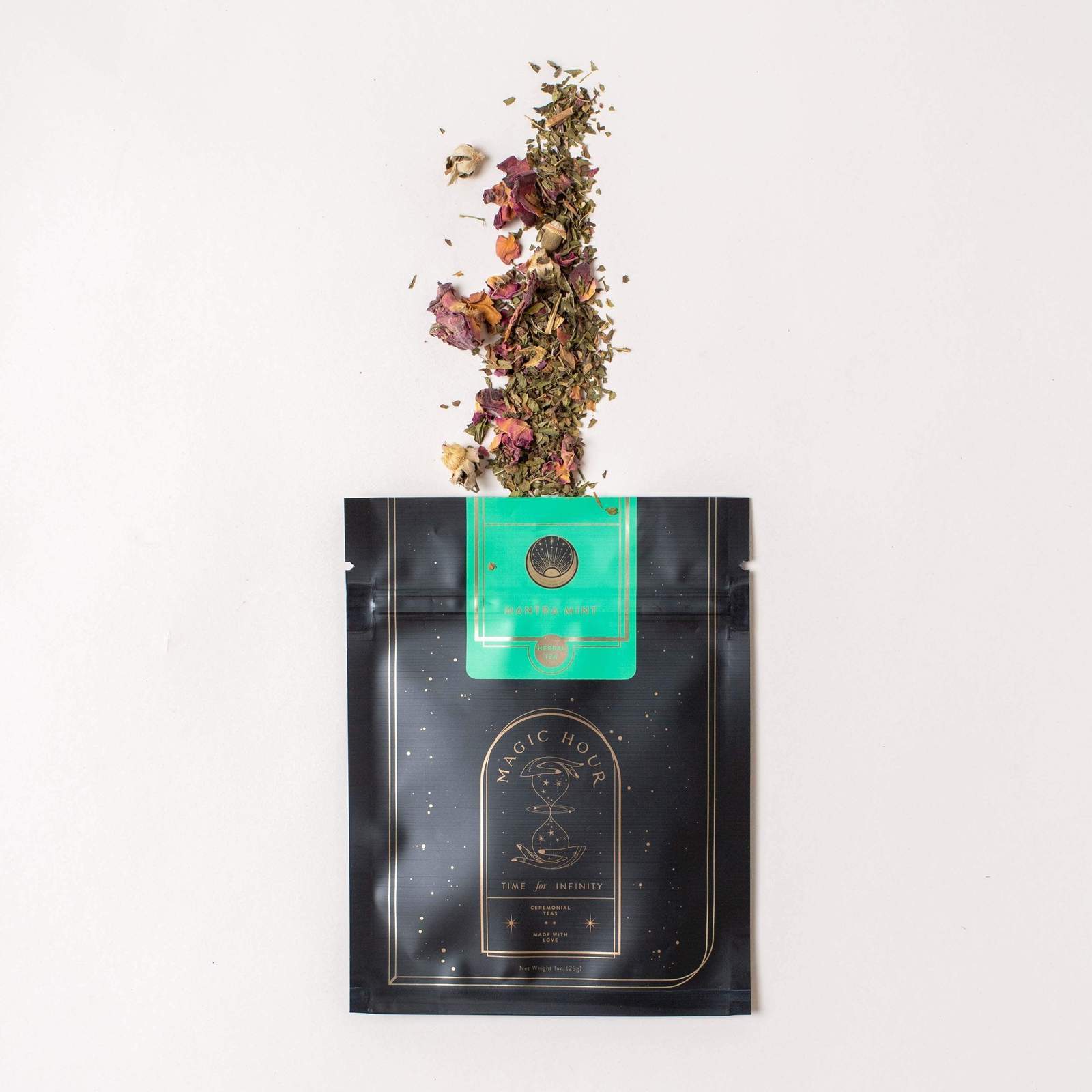 A black pouch with gold detailing labeled "Magic Hour Renew & Relax: Ritual Gift for Mindfulness & Calm" has its top open, with a smaller green packet labeled "Cistus Incanus" partially pulled out. Loose leaf tea, including dried flowers and leaves, has spilled from the packets onto a light surface.