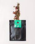 A black pouch with gold detailing labeled "Magic Hour Renew & Relax: Ritual Gift for Mindfulness & Calm" has its top open, with a smaller green packet labeled "Cistus Incanus" partially pulled out. Loose leaf tea, including dried flowers and leaves, has spilled from the packets onto a light surface.