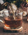 A glass teapot pours Magic Hour Gamma-Aminobutyric Acid (GABA) Oolong Tea into a clear glass cup placed on a wooden coaster. Another cup of tea is in the background, slightly out of focus, enhancing the calm nourishment of the rustic setting with muted tones and blurred flowers.
