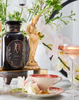 A decorative scene featuring a black jar labeled "Astrology Bundle: Aries, Taurus, Gemini - Magic Hour" adorned with gold designs, flanked by two ornate figurines. Surrounding the jar are elegant flowers, a filled cocktail glass, and a teacup with saucer showcasing Organic Tea. The backdrop is adorned with lush greenery.