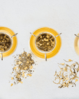 Three glass teacups containing light-colored teas are placed on a white surface. Next to each cup, there are small piles of loose leaf tea and ingredients: chamomile, green tea with flowers, and dried plant roots, representing the Gemstone Bundle with Diamond, Emerald, and Pearl from Magic Hour.