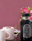 A bottle of Gypsy Rose Black Tea by Magic Hour sits on a round golden tray with a black base. Next to it are two white cups filled with tea, garnished with small rose petals. In the background is a bouquet of pink roses against a mauve wall.