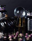 A dark, elegant display features two black jars labeled "Black Tea Magic Sampler Box- Bestselling Organic Black Teas for Energy & Vitality," a black teapot with gold accents, dried rosebuds, a crystal ball on a stand, a crystal cluster, and glassware against a dark backdrop. The scene exudes mystical and luxurious vibes with the allure of Magic Hour.