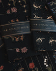 Four packaged Tea Ceremony Towels labeled "Magic Hour" lie on a decorated surface. Each towel features light gold and colored floral and celestial designs against a dark background. Two packages showcase constellations, while the other two highlight flowers.