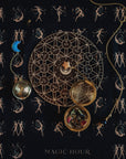 A celestial-themed flat lay showing a zodiac wheel, a locket with herbs and stones, and a crescent moon ornament, all placed on dark fabric adorned with mystical illustrations of mermaids, dancing figures, and stars. The words "Tea Ceremony Towels Bundle" by Magic Hour are at the bottom, evoking the charm of organic tea rituals.