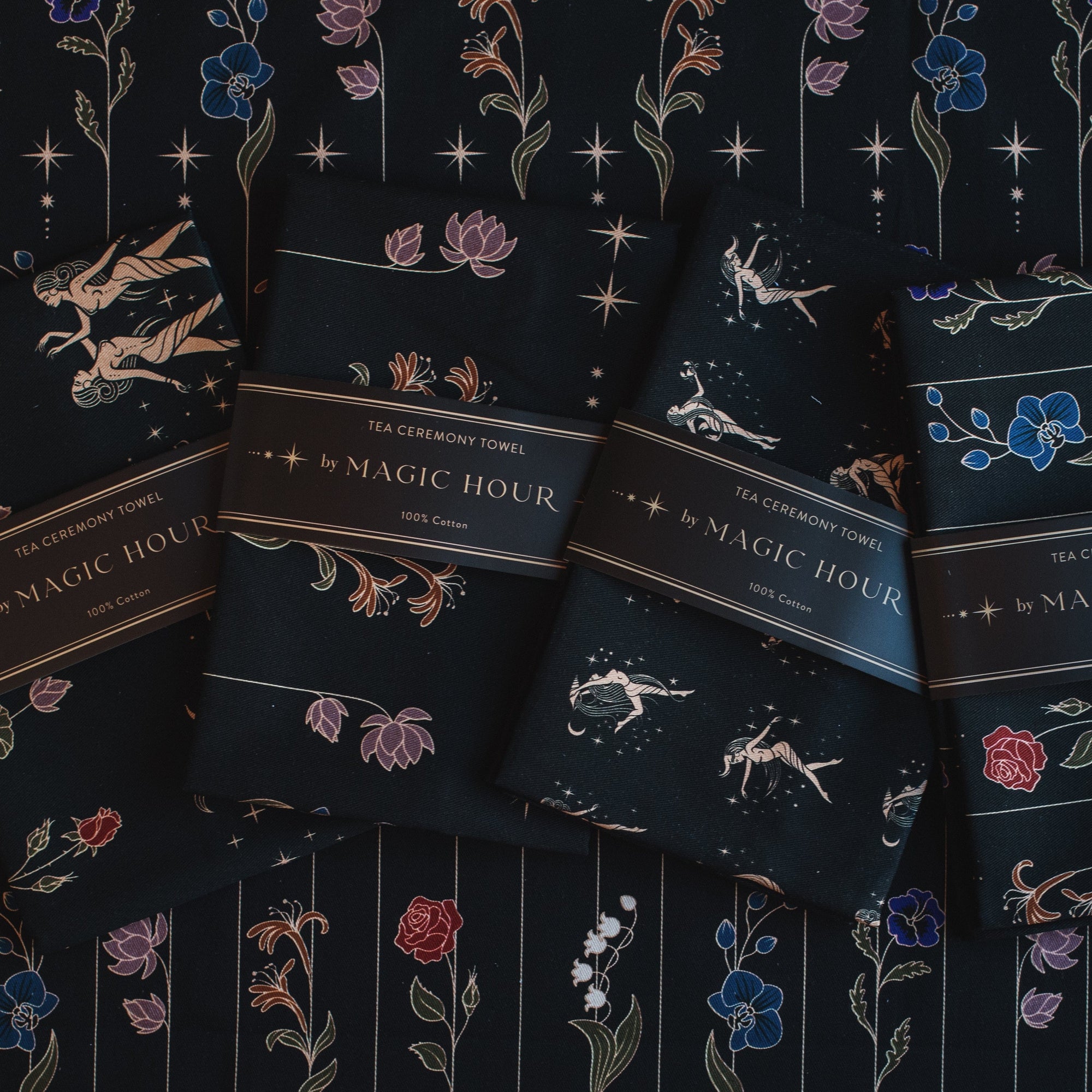 Four Tea Ceremony Towels are displayed on a dark, patterned surface. Each towel is wrapped with a band that reads "Tea Ceremony Towels by Magic Hour." These elegant pieces, perfect for your Magic Hour tea rituals, feature intricate designs of flowers, celestial bodies, and constellations.