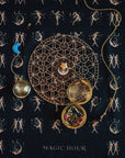 A mystical setup featuring a geometric and lunar phase chart, a gold locket holding various crystals, and a moon-shaped pendant, all set on a celestial-patterned cloth with dancing figures. A steaming cup of loose leaf tea completes the scene. The caption at the bottom reads "Magic Hour Tea Ceremony Towels.