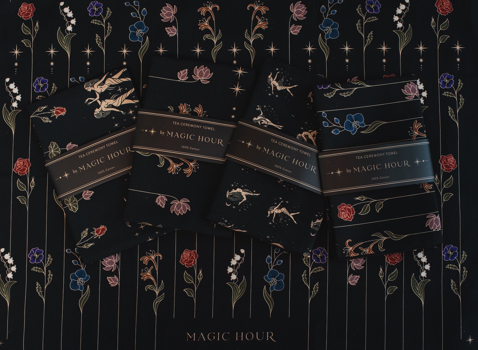 Four dark-colored packets of Magic Hour Tea Ceremony Towels are arranged in a row on a floral-patterned background. The packets feature small illustrations of celestial elements and flowers in shades of gold, white, and blue, capturing the enchanting essence of this organic loose leaf tea.