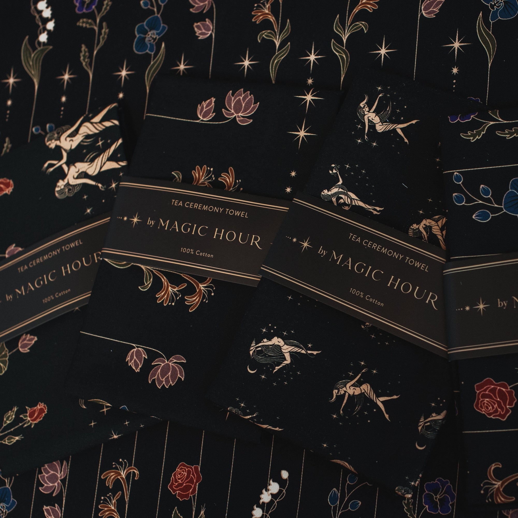 A collection of elegant Tea Ceremony Towels by "Magic Hour," featuring intricate floral and celestial designs in gold, blue, and red on a black background. Each towel is labeled "100% Cotton." They are artistically arranged, showcasing their elaborate patterns and perfect for your loose leaf tea rituals.