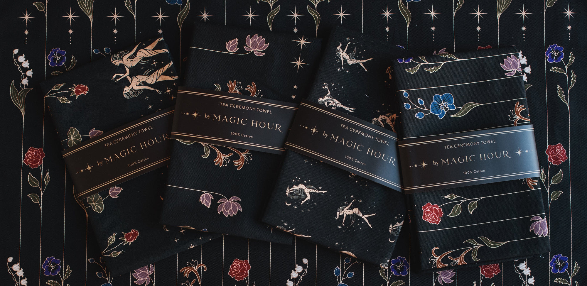 Four Tea Ceremony Towels with intricate designs and labels that read "Magic Hour" are displayed. The designs include flowers, leaves, and celestial elements. They are laid out on a dark, intricately patterned fabric that complements the towels' designs, evoking the essence of organic tea rituals.