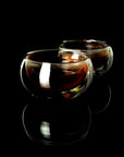 Two glass teacups filled with dark liquid, possibly Sin Eraser™ : Puerh Tea by Magic Hour, are placed on a glossy black surface. The cups have a double-walled design, creating a clear and elegant appearance, with reflections visible beneath them. The background is completely black.