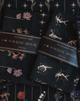 A display of four neatly folded Tea Ceremony Towels Bundle, each wrapped with a label reading "Magic Hour." The towels feature various floral and celestial designs in vibrant colors, placed on a fabric backdrop adorned with intricate floral and star patterns.