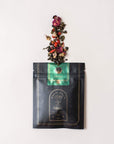 A black and green Magic Hour Renew & Relax: Ritual Gift for Mindfulness & Calm package with intricate gold detailing is laying on a beige surface. The package is open at the top, and a mix of organic loose leaf tea leaves and colorful flower petals is spilling out in a visually appealing arrangement.