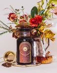 A display featuring a black and gold jar of "Queen of the South: Delicious Cocoa Detox Tea" by Magic Hour, a golden tea strainer with loose tea leaves, a mug of organic tea with cinnamon, a metal pitcher of milk being poured, and a vibrant floral arrangement in the background.