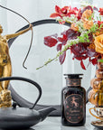 A decorative setup featuring a gold archer statue, a black teapot, a vase with vibrant red, orange, and white flowers, and a black jar labeled "Sagittarius Tea of Good Fortune & Abundance Magic Hour". A teacup and saucer are placed beside the jar, all on a light surface.