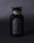 A dark glass bottle with a black label marked "Magic Hour" and illustrations, boasting the text "Time & Infinity." The bottle, reminiscent of Magic Hour's refined tea blend Ti Quan Yin Oolong: Tea of Patient Compassion, has an ornate design and a stopper on top. The background is a gradient of dark gray shades.