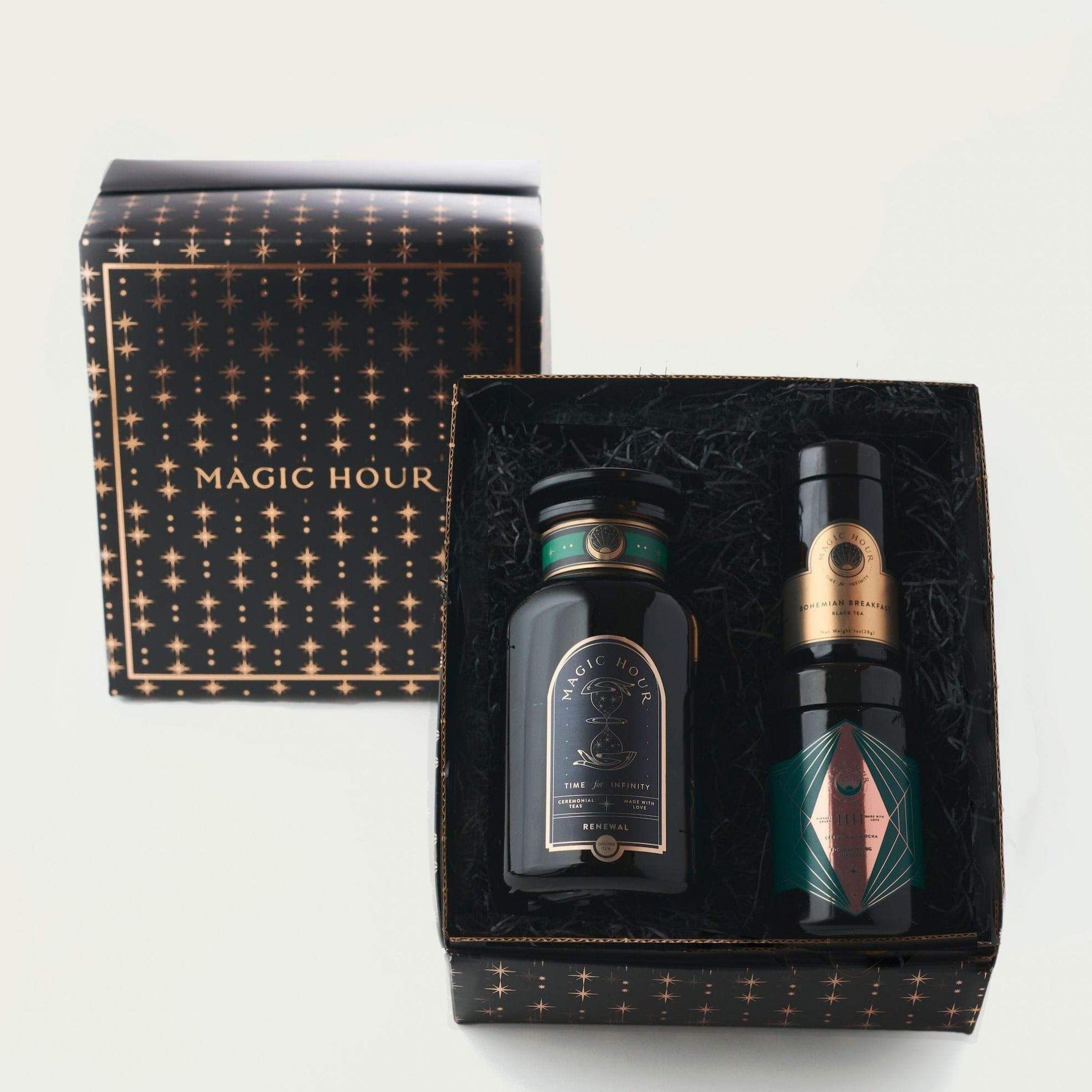 A gift box set labeled "Tea Connoisseur Collection" from Magic Hour contains three items. One is a candle in a black jar with gold accents, another is a small cylindrical container of loose leaf tea with a black lid, and the third item is similar in shape with a part-green, geometric design.