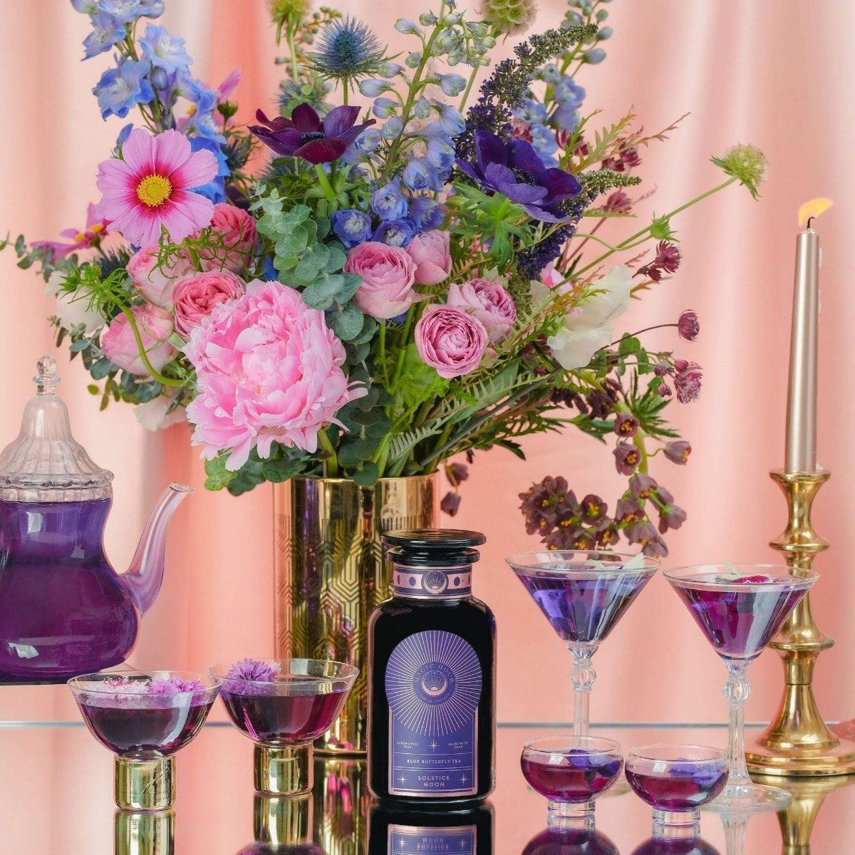 A vibrant floral arrangement in a gold vase is accompanied by a towering lit candle, multiple glasses of the purple Solstice Moon™ drink, and a clear teapot containing an organic tea blend. The scene is set against a soft pink backdrop.