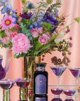 A vibrant floral arrangement in a gold vase is accompanied by a towering lit candle, multiple glasses of the purple Solstice Moon™ drink, and a clear teapot containing an organic tea blend. The scene is set against a soft pink backdrop.