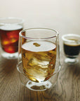 Three double-walled glass mugs are shown on a wooden table. One contains an amber-colored drink with ice cubes, another has a dark brown beverage, and the third, smaller mug holds a dark espresso shot. The amber drink is a refreshing blend of organic tea that perfectly complements the setting in the Kinto Double-Walled Iced Tea Glass: 350mL by Magic Hour.