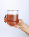 A hand is holding a Kinto Double-Walled Iced Tea Glass: 350mL filled with a light brown liquid against a plain white background. The glass is rounded and transparent, showcasing the Magic Hour Organic Tea inside clearly.