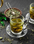 Two glass cups of Magic Hour Mao Jian (Emerald) Green Tea of Good Fortune on saucers sit on a dark surface, surrounded by loose green tea leaves. A wooden bowl filled with more organic tea leaves and a tea strainer are nearby. Green tea leaves and a brass teapot add to the cozy, relaxed atmosphere.